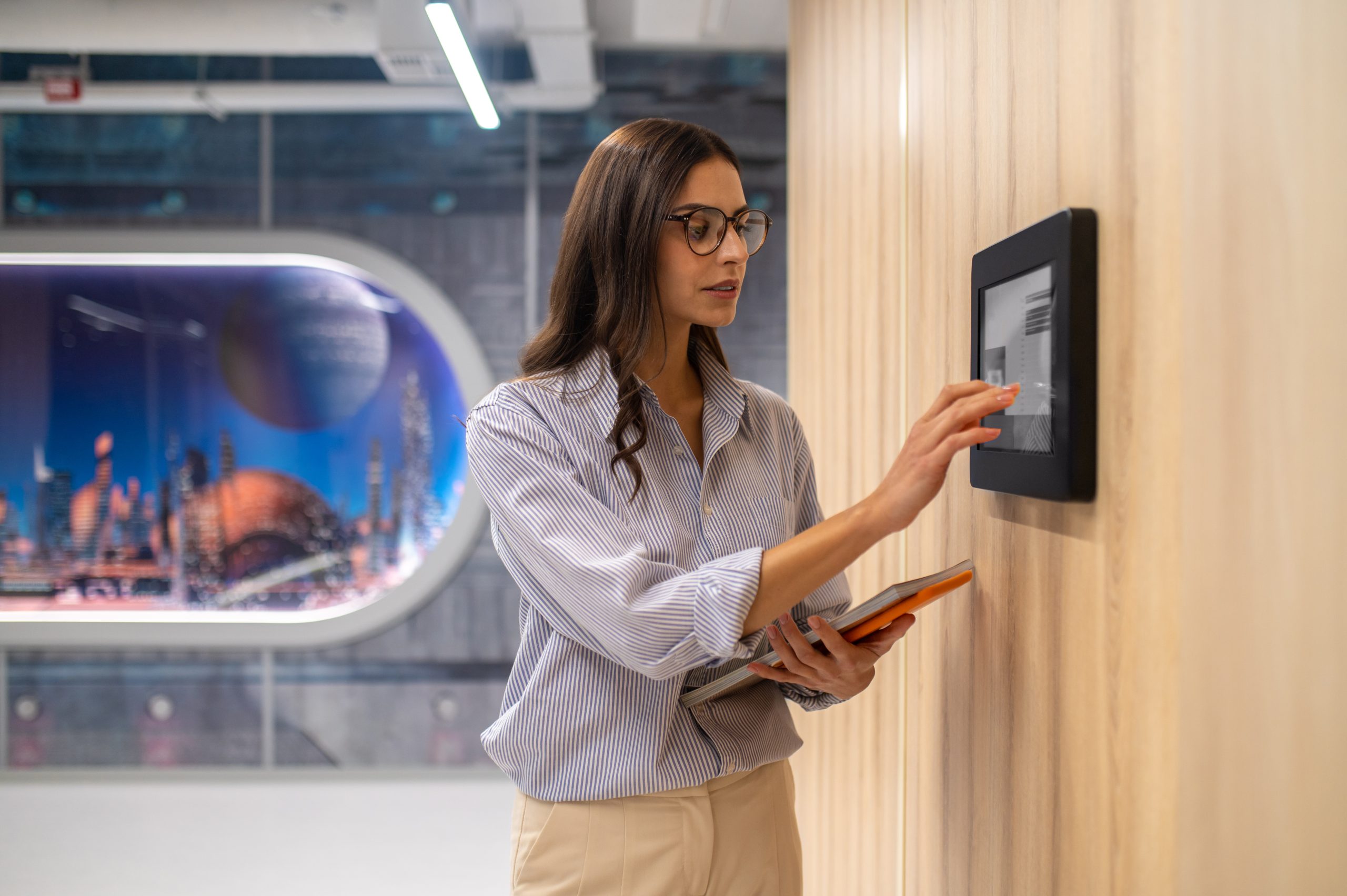 Control panel. Focused elegant woman in glasses with folder touching with hand control panel on wall in illuminated corridor of school